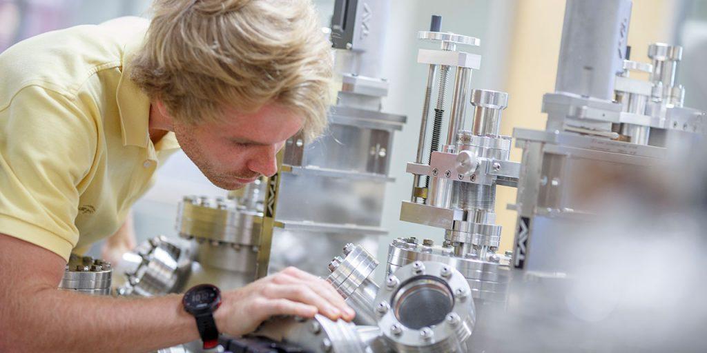 An engineering student in a lab peers into a small window in a large metal device. 