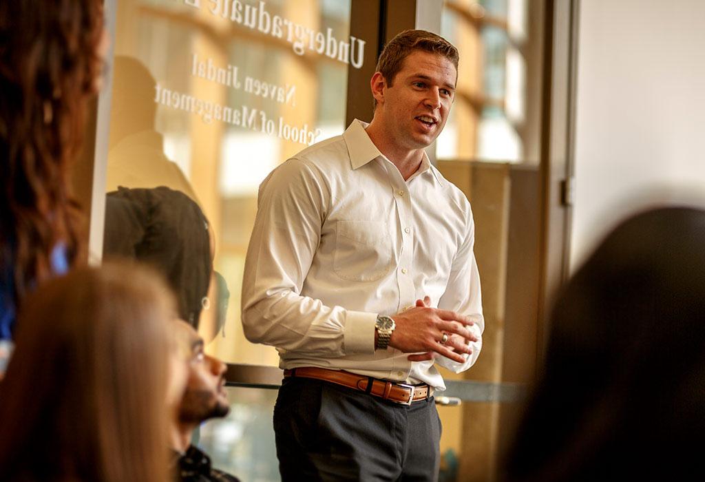 A person speaks to several others in front of a glass door that reads Undergraduate and Naveen Jindal School of Management. 