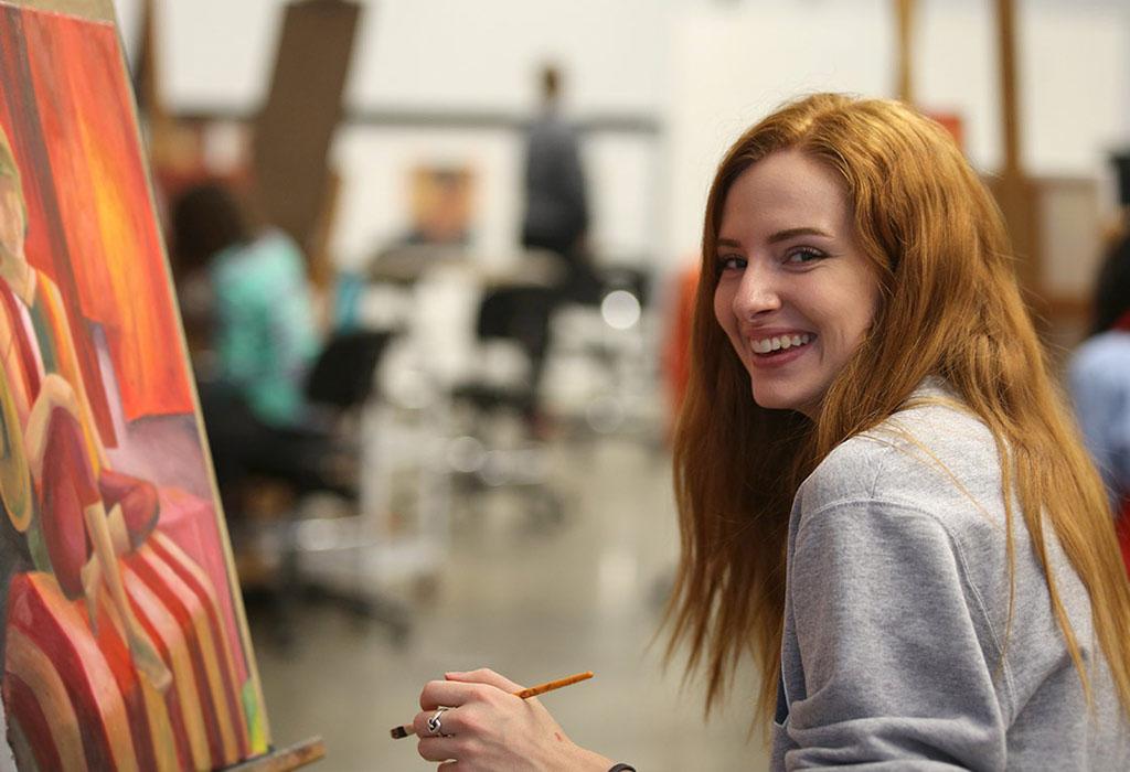 A student seated at a painting with brush in hand. They've paused their work to smile at the camera. 
