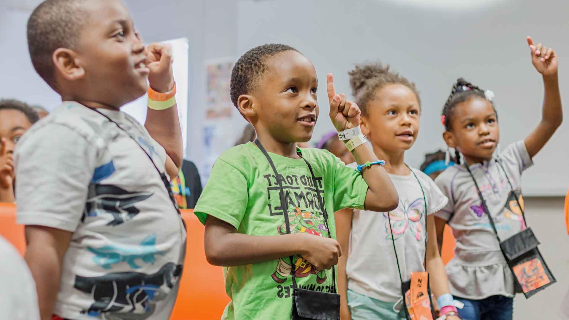 Four little kids at a summer camp. They're wearing brightly colored shirts and bracelets and several are raising a hand and index finger. 