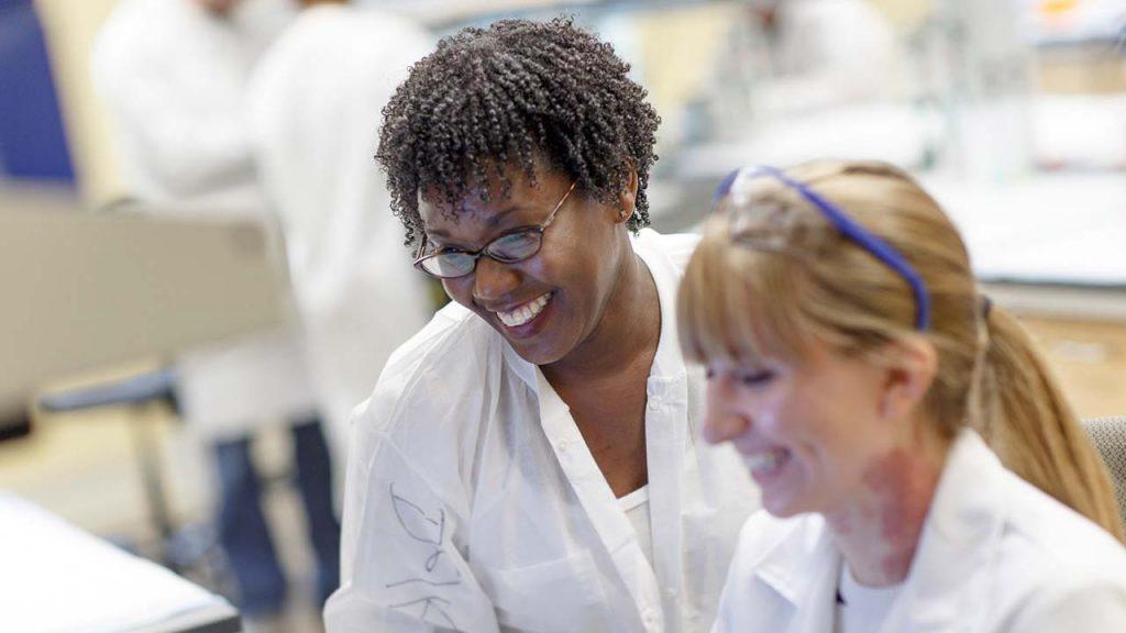 Two faculty members in lab coats, with other researchers out of focus in background. 